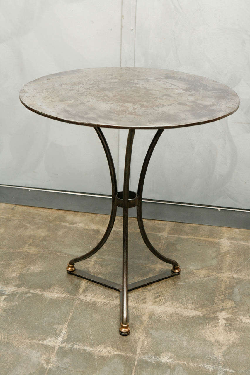This beautifully designed french iron garden table has a rustic finished table top and polished brass details at the base of the feet. The three splayed Iron legs are nicely shaped with a ring that joins the three stems. This piece works well as a