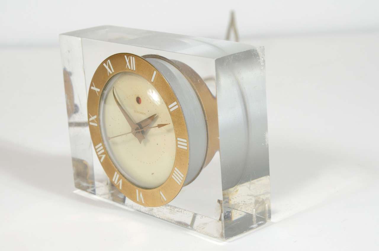 This beautiful clock features a Brass relief dial with Enamel detailing with a floating lucite body resting on stylized Deco inspired rests.It has an alarm as well.