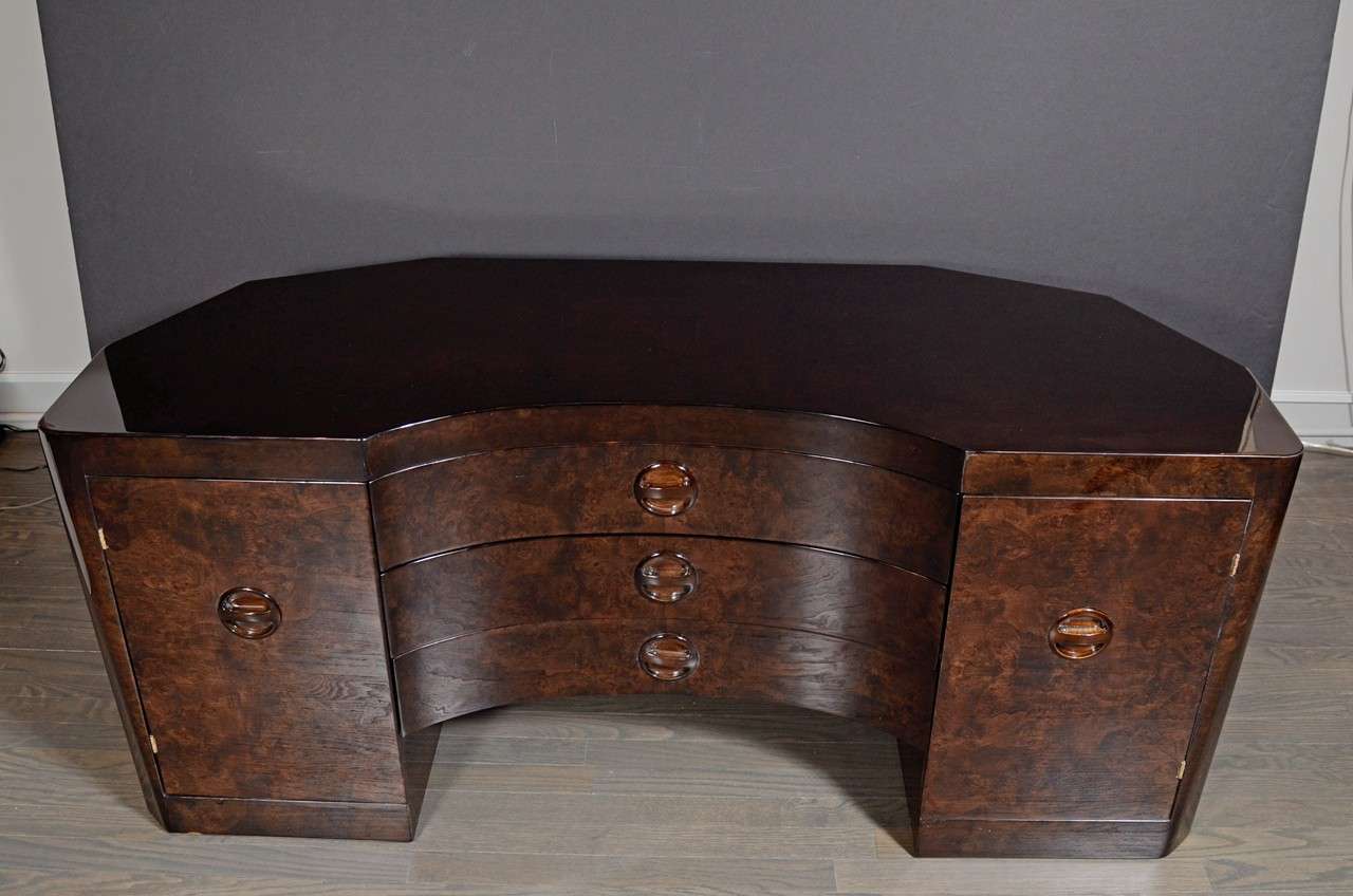 This gorgeous Machine Age Art Deco vanity was realized in the United States, circa 1935. It features has a wraparound mirror realized in bookmatched and ebonized walnut with machine age pulls. The piece offers two cabinets with shelves and three