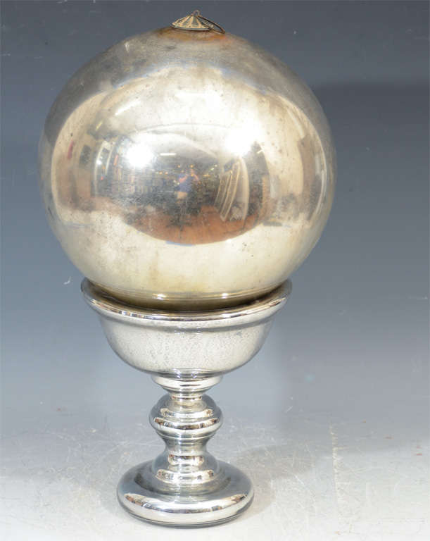 An antique Mercury glass witch ball on a modern chrome pedestal. There is a hanging hook on the crown of the piece.

Traditionally witching balls were used to ward off evil and were placed on porches or in gardens.