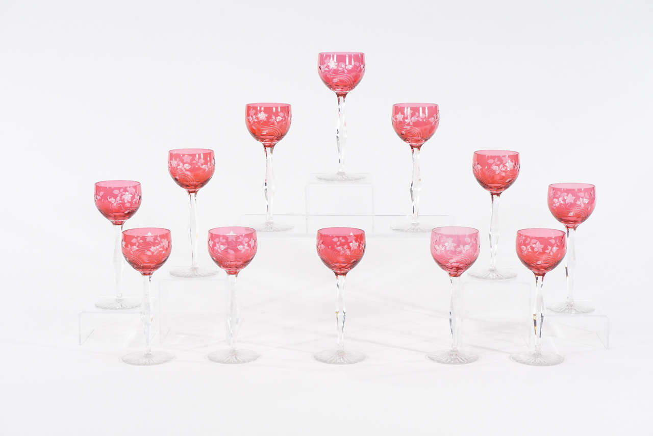 This rare set of 12 Baccarat goblets truly embody the Art Nouveau aesthetic, featuring a cranberry crystal bowl sitting atop a tall and elegant panel cut stem. The goblet is cut to clear in a swirling intaglio motif with loosely floating flowers and