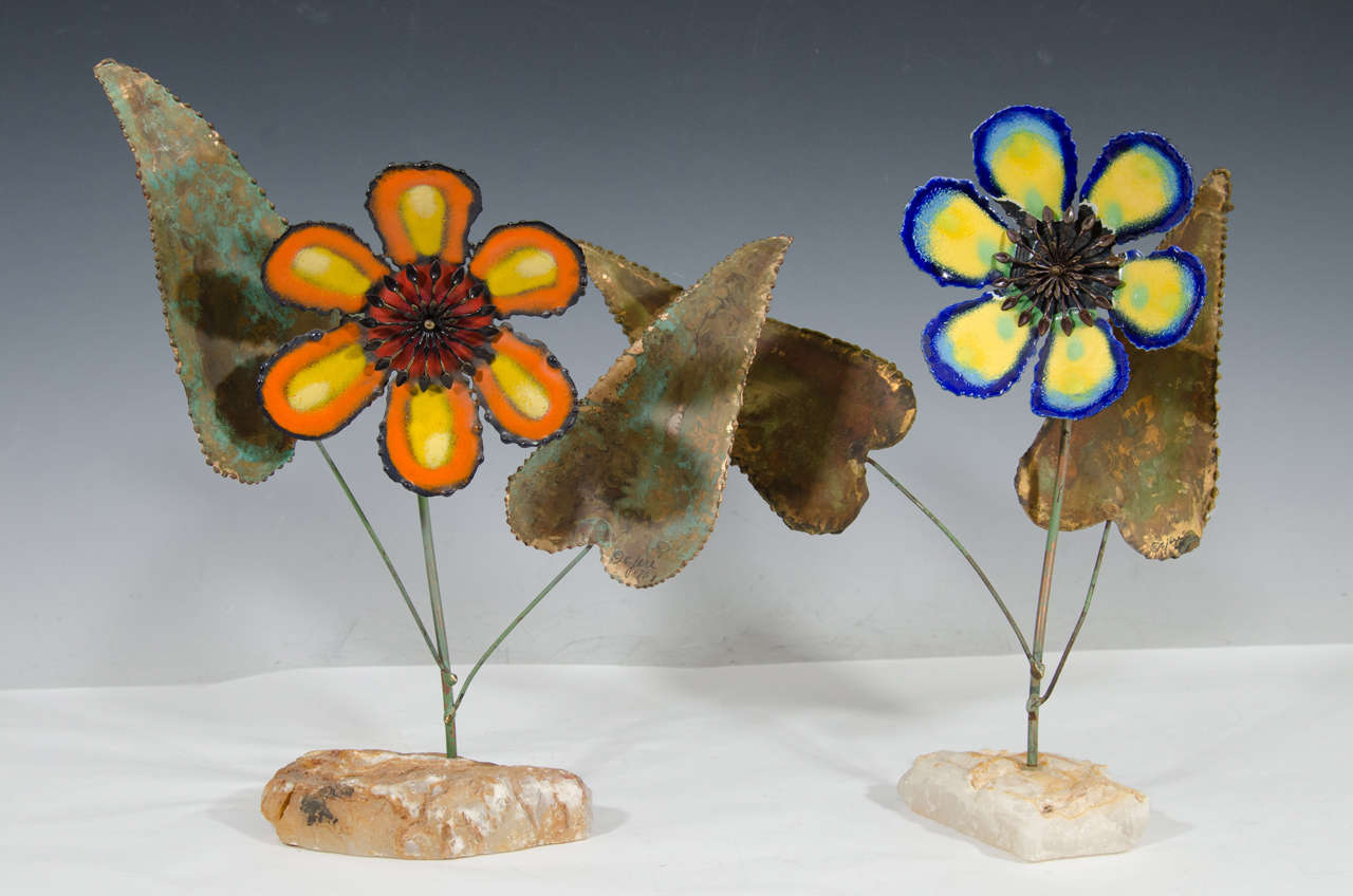 A vintage set of two flower sculptures on quartz bases with metal leaves by Curtis Jere.  One enamel flower is blue, yellow, and green.  The other flower is orange, yellow, and red. Signed C. Jere. Good vintage condition with age appropriate patina.