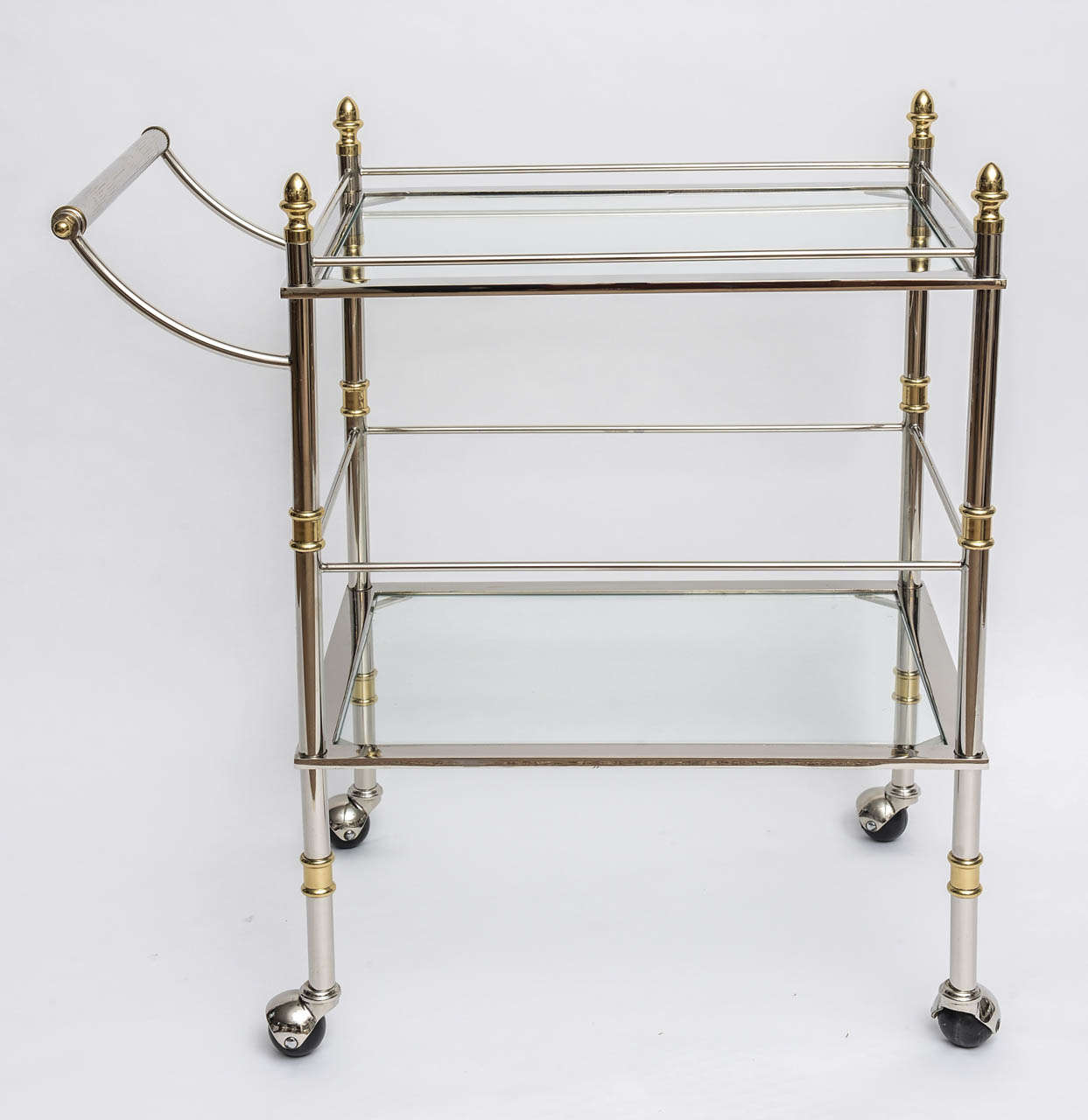 Gorgeous rolling bar or serving cart with two tiers of glass shelves. The top shelf has the fencing around it and the frame is in polished nickel adorned with brass accents including the acorn top finials. The wheels roll perfectly with ease and are