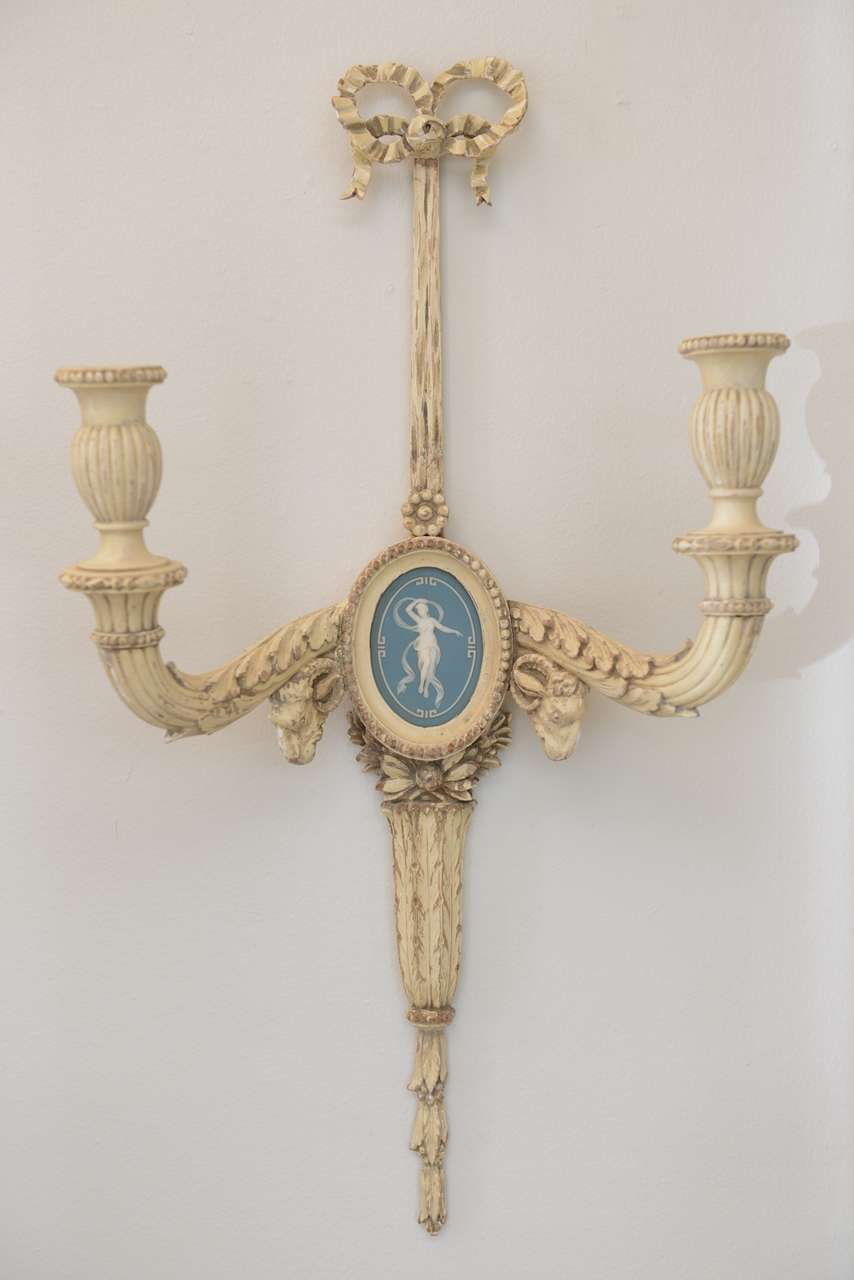 Opposing pair of sconces, of wood, having distressed painted finish, each surmounted by intricately carved bow; two scrolling candlearms supported by rams' heads, centered with oval blue Wedgewood bisque plaque and terminating in foliate and laurel