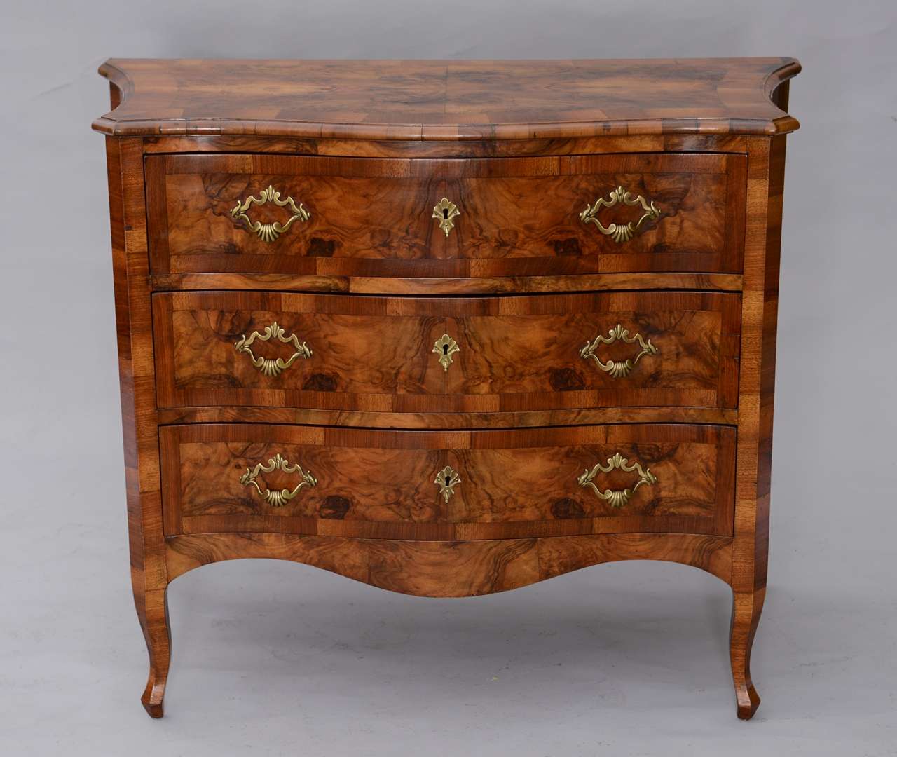 Commode, having an olivewood burl veneer, its shaped and molded top, on conforming case, over three stacked drawers, bowed apron raised on cabriole legs.