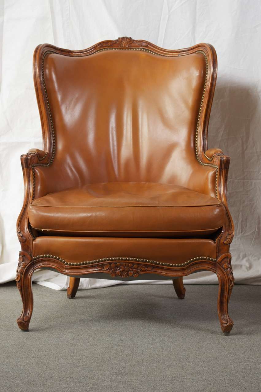 This pair of Louis 15th style bergere chairs features nail head trim and slightly distressed carmel colored leather. The high back with a slight barrel shape gives them a wing chair feel , perfect of a library or at the fireplace. Carved floral