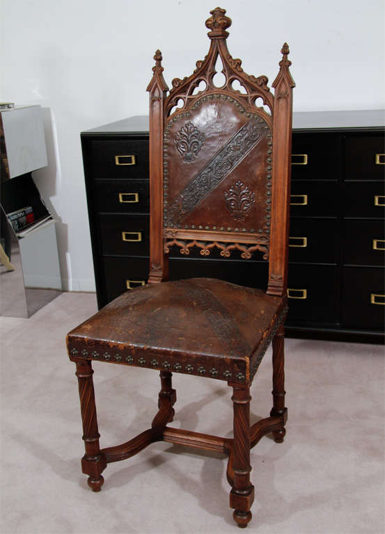 A pair of gothic chairs in hand carved wood with tooled leather backs and seats. The seatbacks have a stylized vegetal motif and the seats and backs have cruciform nail head detailing.