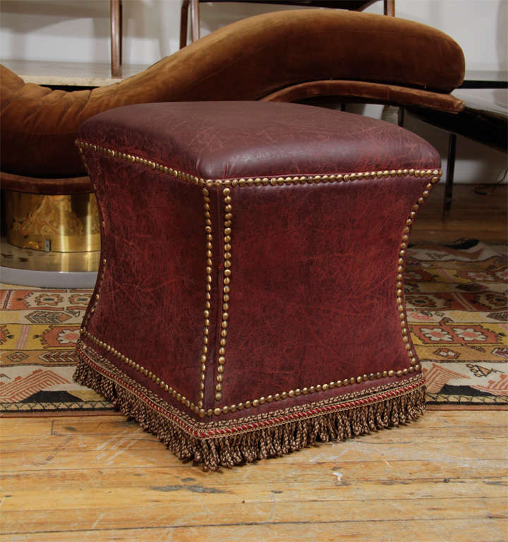 Deep purple distressed leather ottoman or foot stool. The piece has brass nailhead detailing as well as a rope fringe skirt along the bottom. Leather upholstery over wood frame.