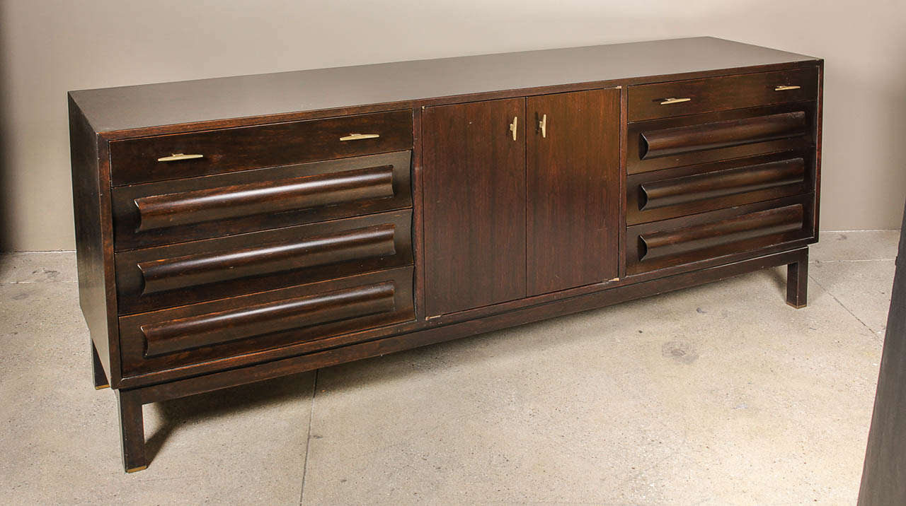 Dark walnut cabinet standing on bronze-tipped chamfered legs. Four drawers at each end form a rhythmic pattern of concave fronts with convex bar pulls. Bronze pulls on upper drawers anc central cabinet, which has one adjustable shelf. Piece is