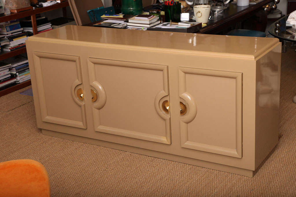 Midcentury sideboard credenza in the style of Paul Laszlo exquisitely lacquered in this rich creamy sand tone. Accented with the cabinets vintage 24-karat gold-plated brass demilune hardware. Four interior drawers to center door and shelving to