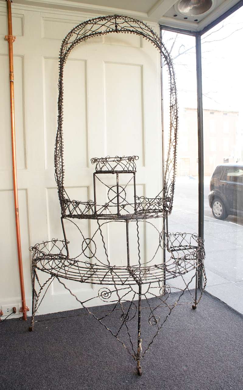 Three tiered wire plant stand, comprised entirely of twisted iron wire. The arched top retains a hook for additional hanging plant. Elaborate, whimsical design. Retains the original wooden casters for mobility.