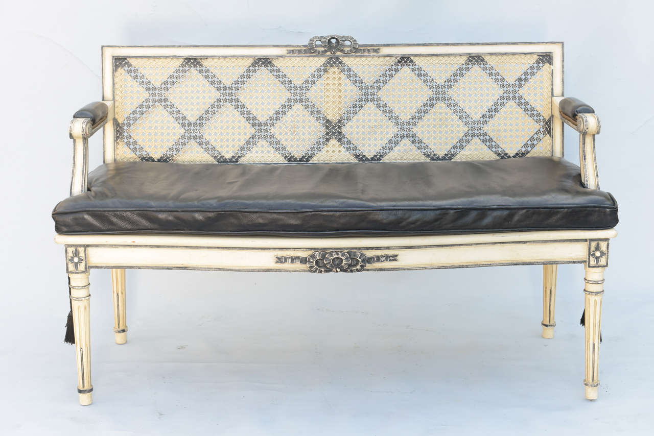 Caned settee, having a painted finish, its fielded crestrail centered with ribbon carving, inset back painted with crosshatch motif, loose cushion of black leather, similarly carved seatrail raised on round tapering fluted legs.

Stock ID: D3117