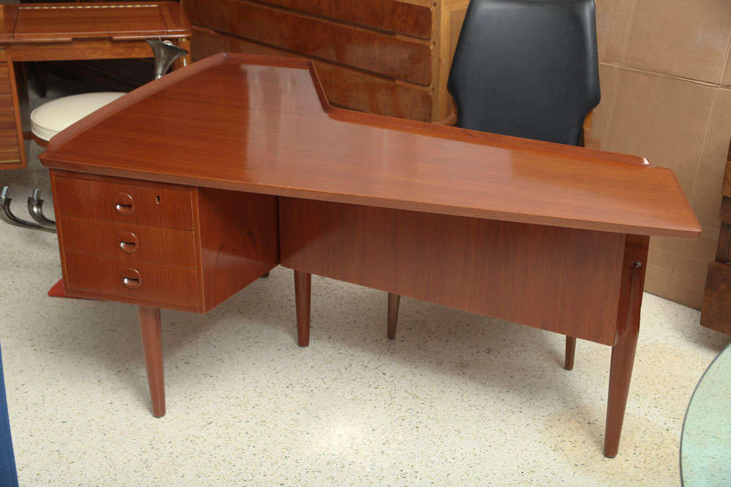 A boomerang shaped desk of teak, features a front work area with three self pull front drawers. The rear has a locking cabinet on the right and a single sliding door over a double storage area on the left. A lip runs around the sides and back ledge.