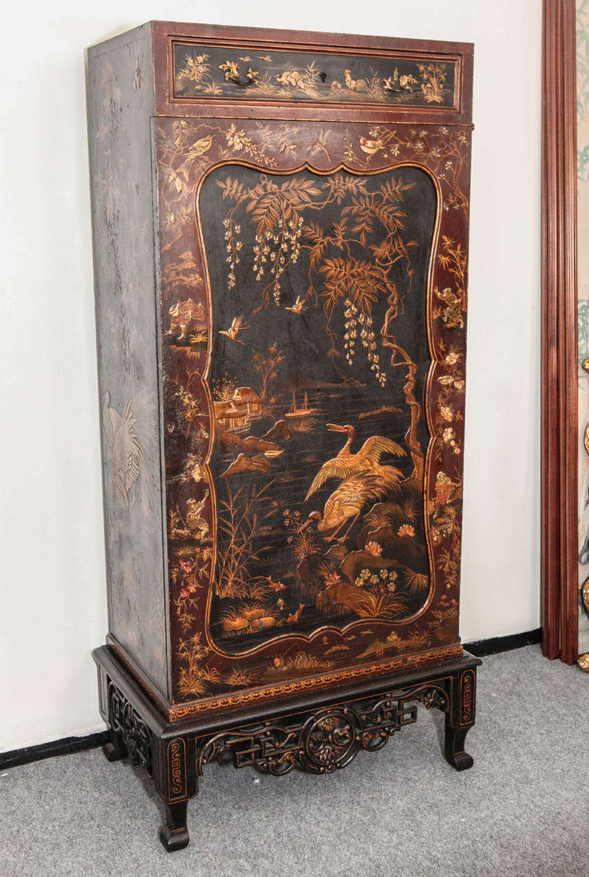 Detailed, hand painted, chinoiserie cabinet featuring an inset scene of crane at a lakeside set on an elaborately carved stand. The interior features seven hidden drawers.