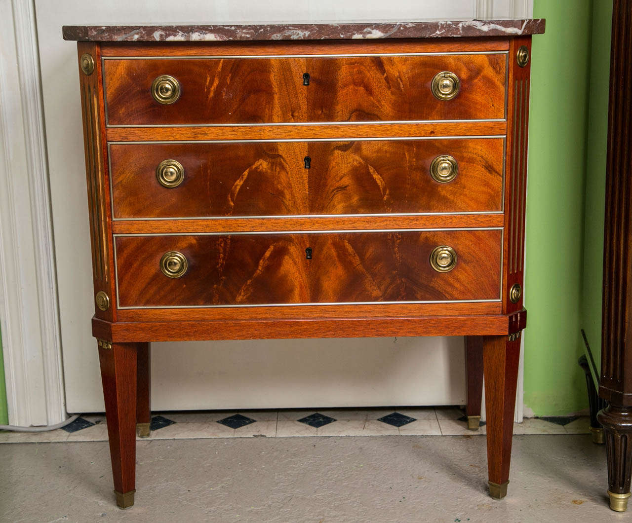 A Sweet Three Drawer Crotch Mahogany Commode by Jansen. This commode / nightstand by Maison Jansen having long legs mounted in bronze decorations leading to fluted bronze corners with bronze circular decorations on top and bottom. The center three