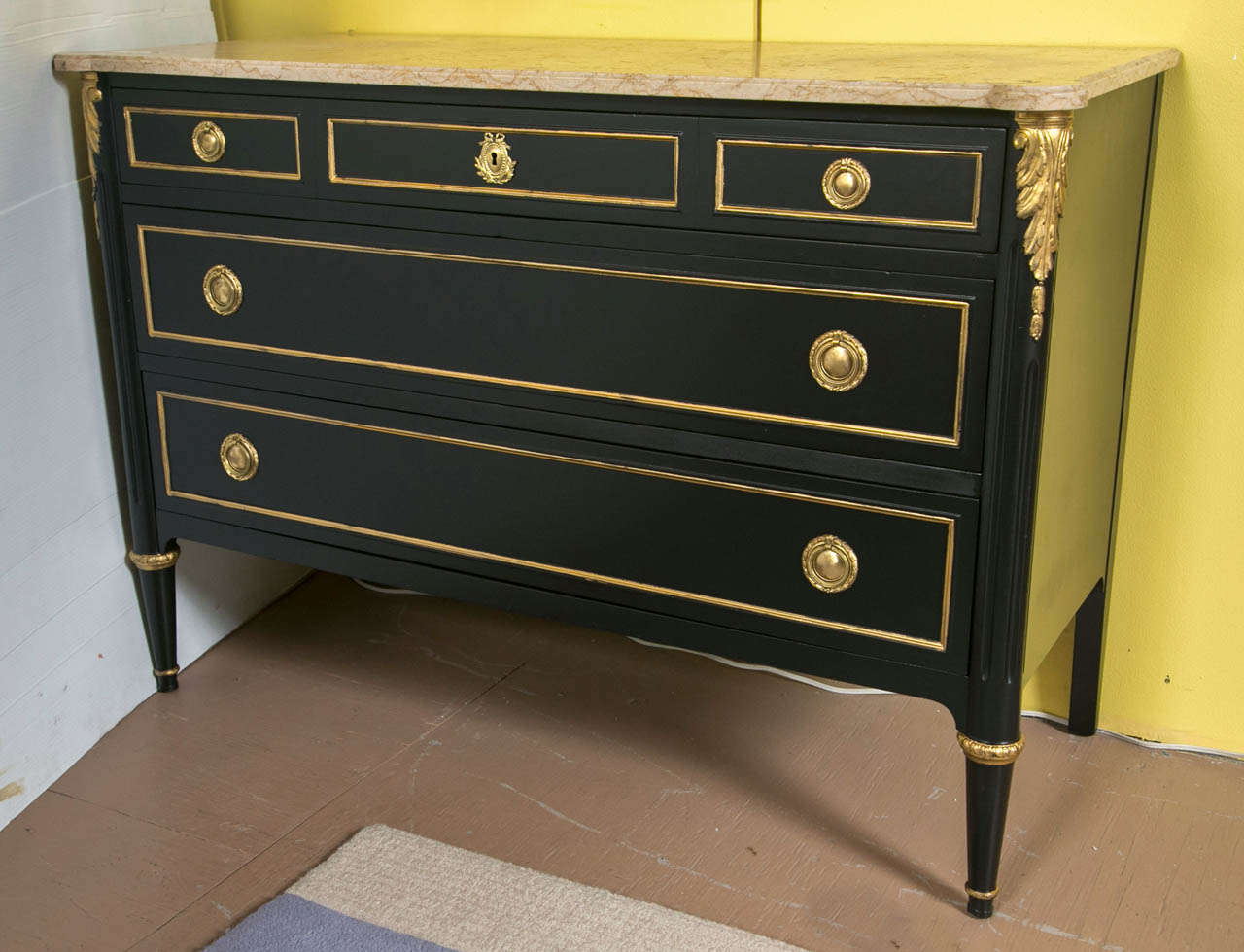 An ebonized marble-top commode by Maison Jansen. Tapering bronze caped feet supporting a group of three drawers. The secondary woods of solid oak. The exterior of a fine ebony finish with gilt gold hi lights and bronze mounts. The top of a beige,