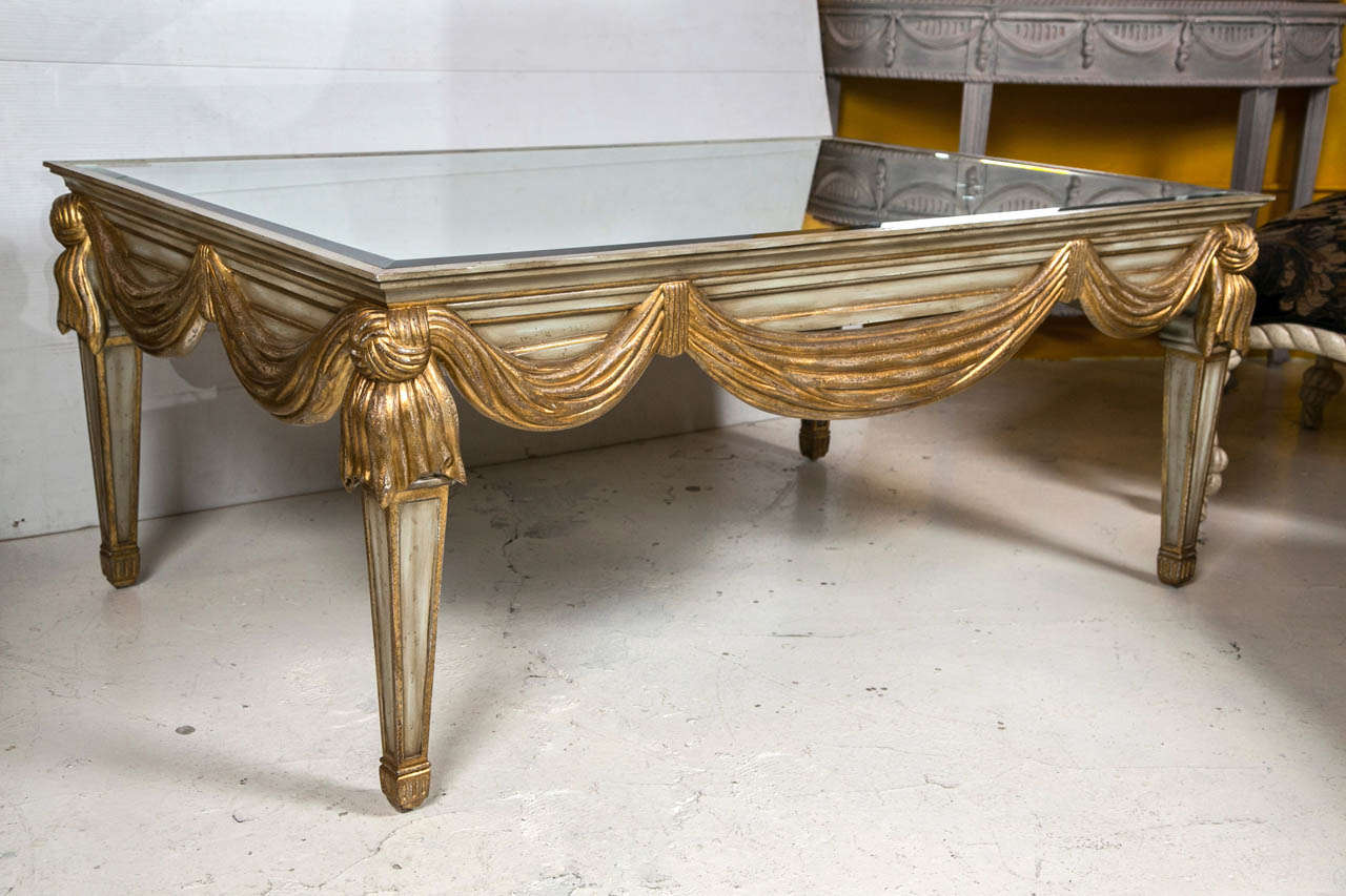 A Ribbon and Tassel Form Decorated Coffee Table in the style of Dorothy Draper. Parcel gilt gold and paint decorated in the Hollywood Regency style base supporting a beveled mirror top.