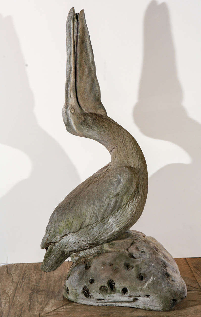 Stone sculpture of a pelican sitting on a rock with it's beak slightly open and pointed upward.  There is an internal  pipe that runs from the base of the sculpture to the tip of the beak.   Gorgeous patina with greys, cremes, and purple tones.