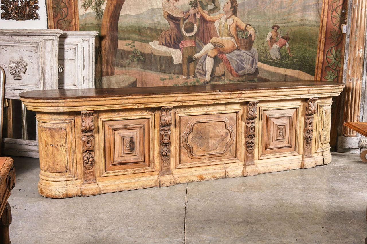 This long oval bistro bar from Spain is absolutely fantastic!  It has been made from stripped European pine and walnut wood. The top is stepped out, and on the front there are four foliate and fruit decorated pilasters.  These separate two raised,