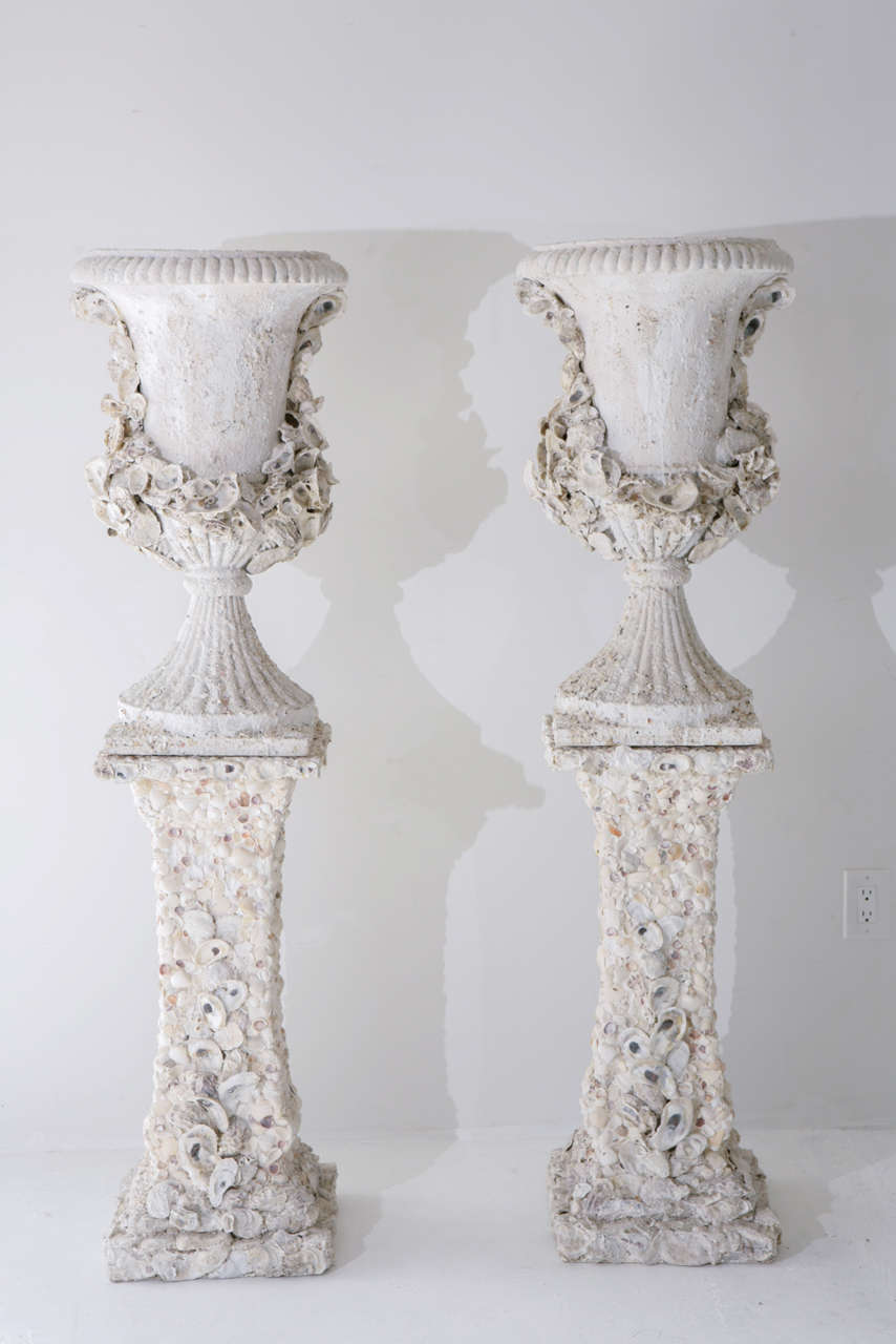 An unusual pair of grotto style cast plaster urns set atop pedestals. The pedestals are completely embellished with seashells and the urns are laureled and dusted with shells. The urns and pedestals are not attached. The urn measures 29.63