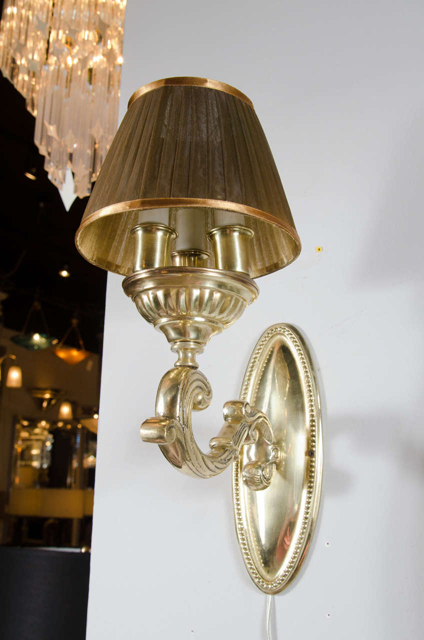 This elegant 1940s Hollywood Regency wall sconce features a brass scroll design with beaded detailing on the base and a flemish scroll arm. It takes three European candelabra base bulbs (max 225 watts) and includes a pleated organza umber shade. It