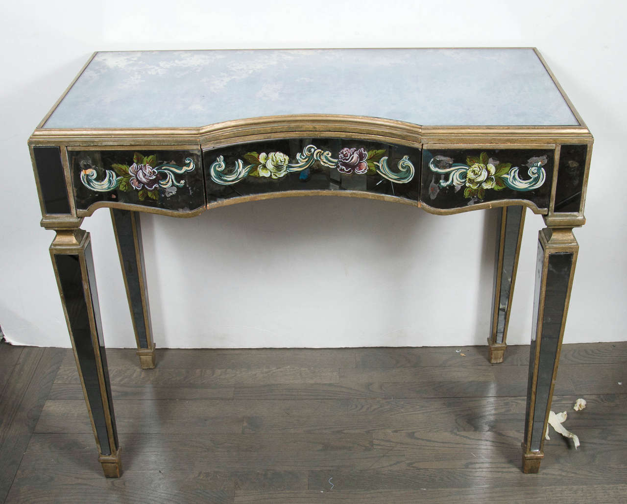 This lustrous 1940s Hollywood Regency console or vanity is elegant in it's design. It features smoked mirror, gilt detailing, as well as the beautiful hand-painted floral and scroll églomisé design, adding to its elegance. It is fitted with three