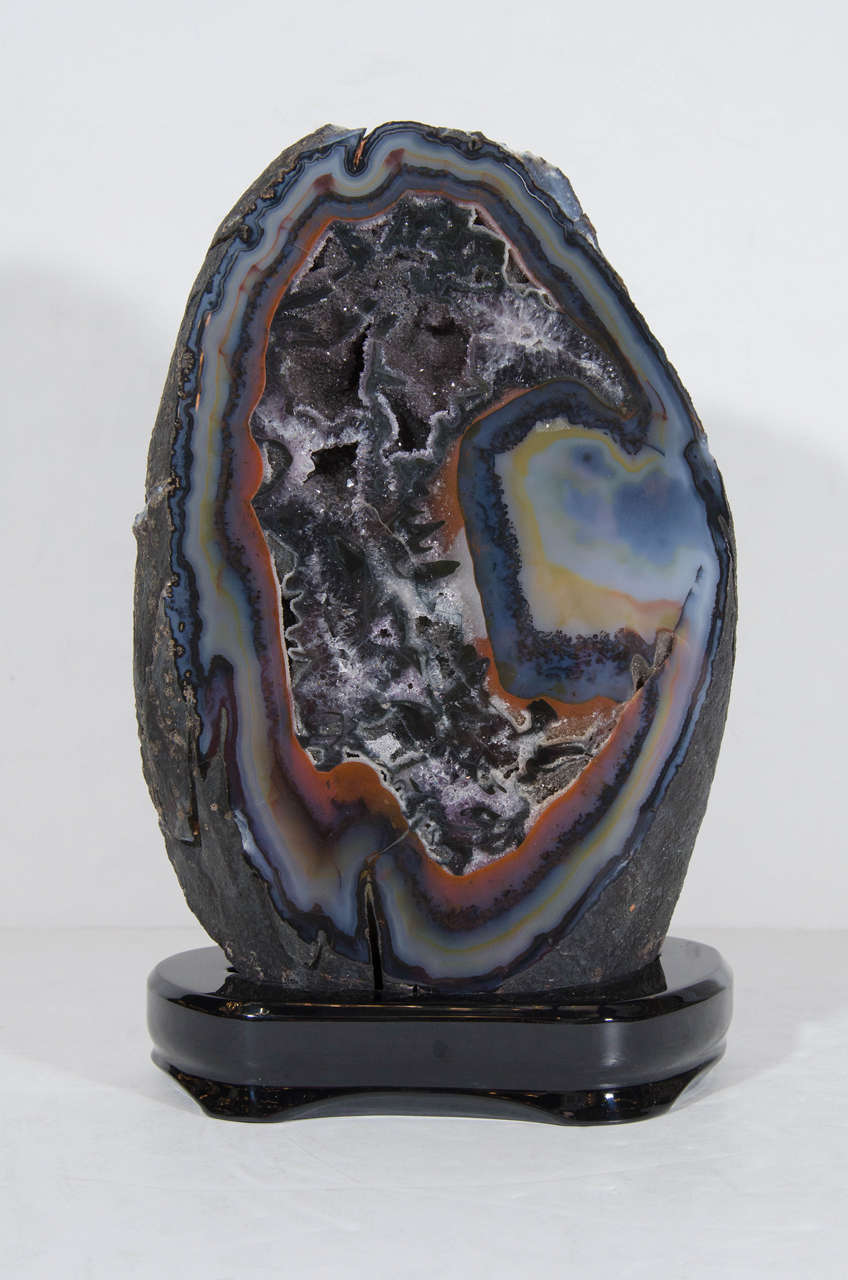 This impressive sliced geode mineral specimen features a natural shape that is trimmed in hues of deep blues, grey, burnt and smokey amber with an interior shell that glitters with smokey crystal. A true eye catcher for any decor and is mounted on a