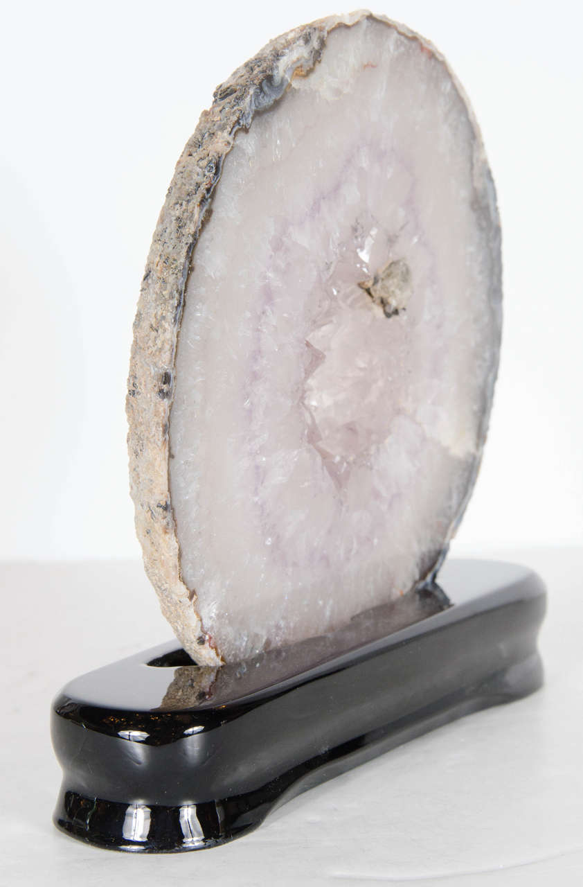 This sliced geode crystal specimen features a circular shape with hues in lavender and greys and pearl, and is mounted on an ebonized walnut base.