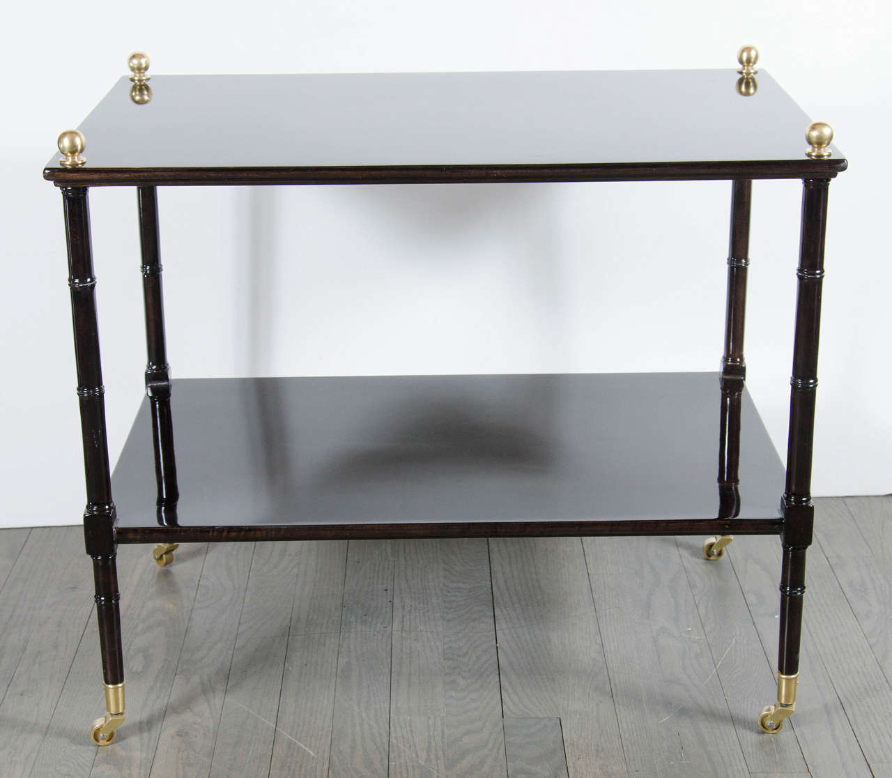 This refined Mid-Century Modern two-tier occasional table was realized in the United States, circa 1960. It features stylized and banded balustrade form legs and two rectangular tops in ebonized walnut with a custom glass top, as well as brass ball