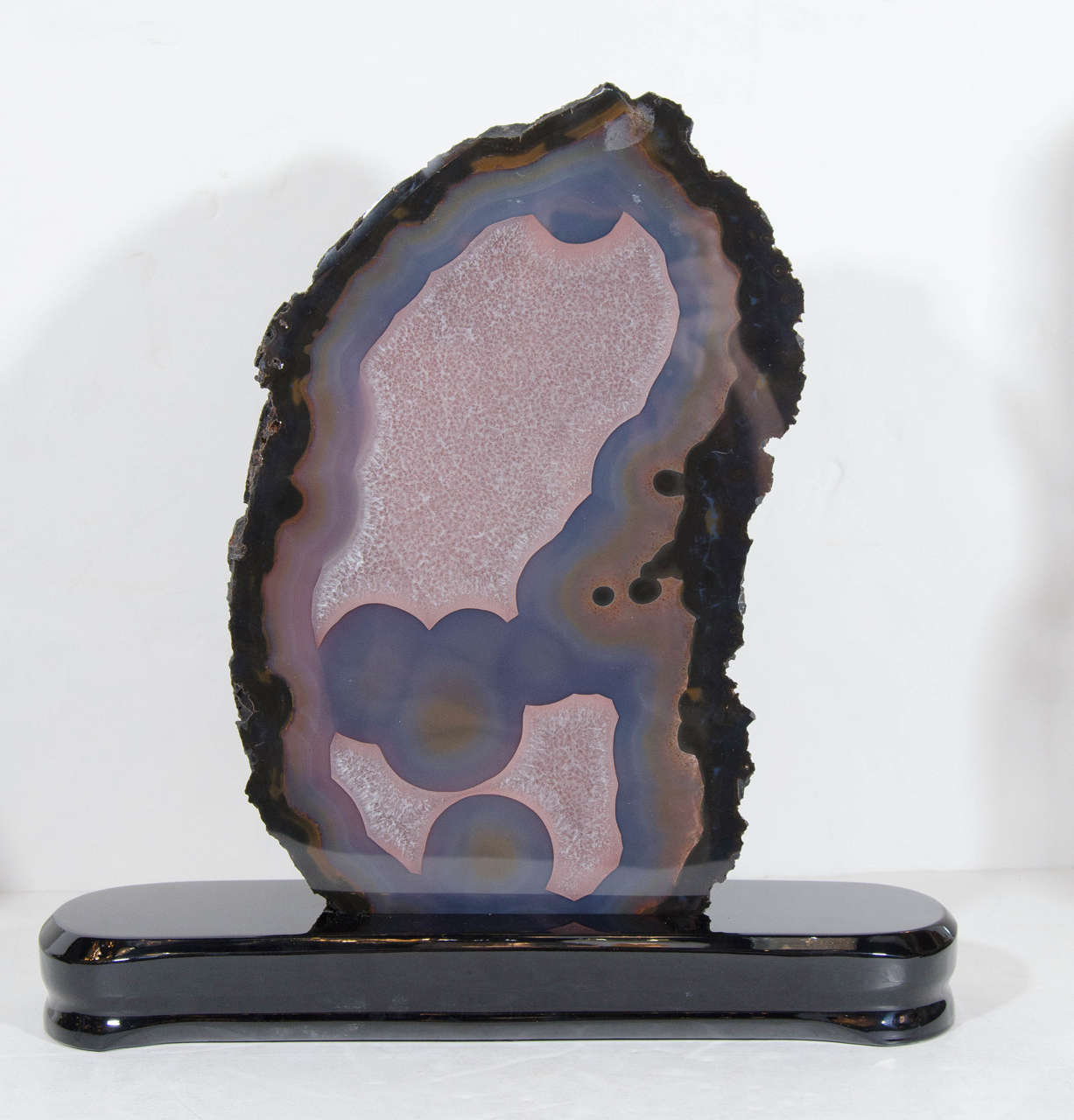 This impressive sliced geode mineral specimen features a natural shape that is trimmed in hues of deep blues, lavender, burnt and smokey umber all amalgamated on a smooth front face. A true eye catcher for any decor and is mounted on a ebonized