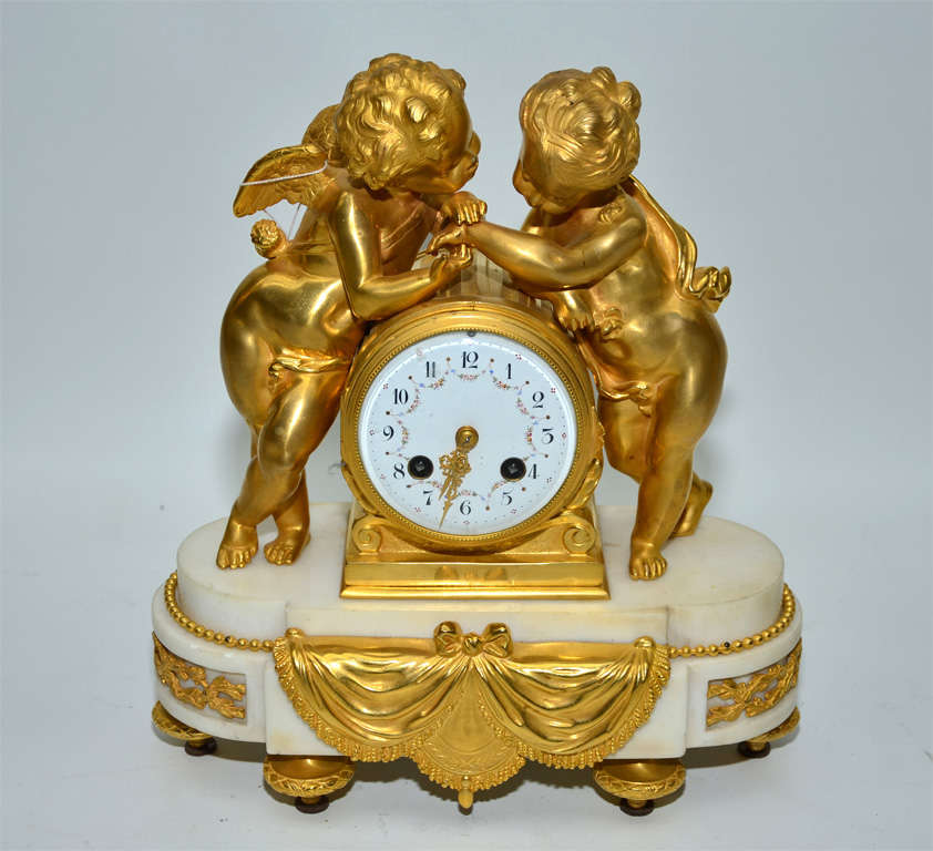 Golden bronze cherubs, surrounding a clock on a white Carrare marble base decorated with golden bronze in  shape of draperie.The  original dial in enamel is decorated with flowers