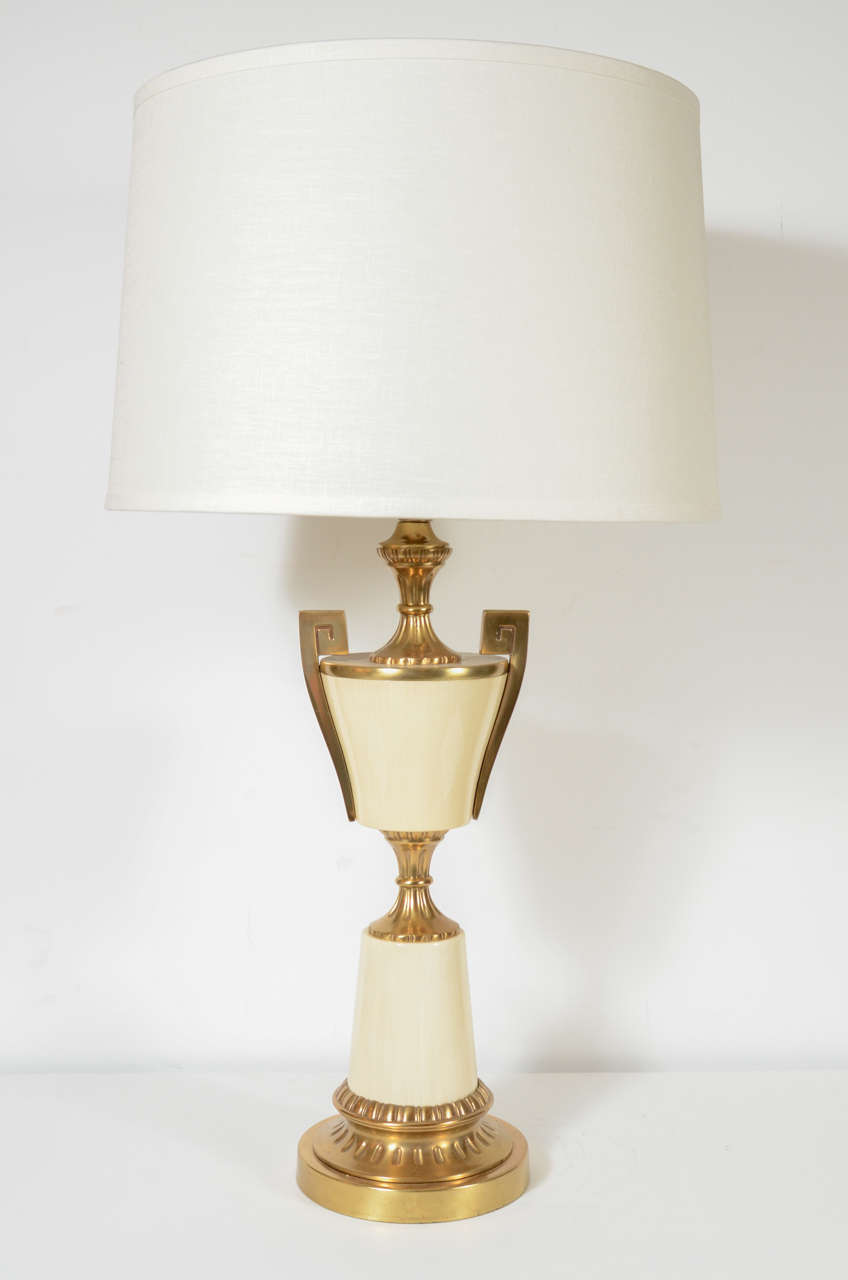 This elegant pair of Mid Century Modern table lamps were realized in the United States circa 1950. They feature cream enamel hour glass form bodies with greek key motifs flanking their shoulders as well as a reeded central embellishment that