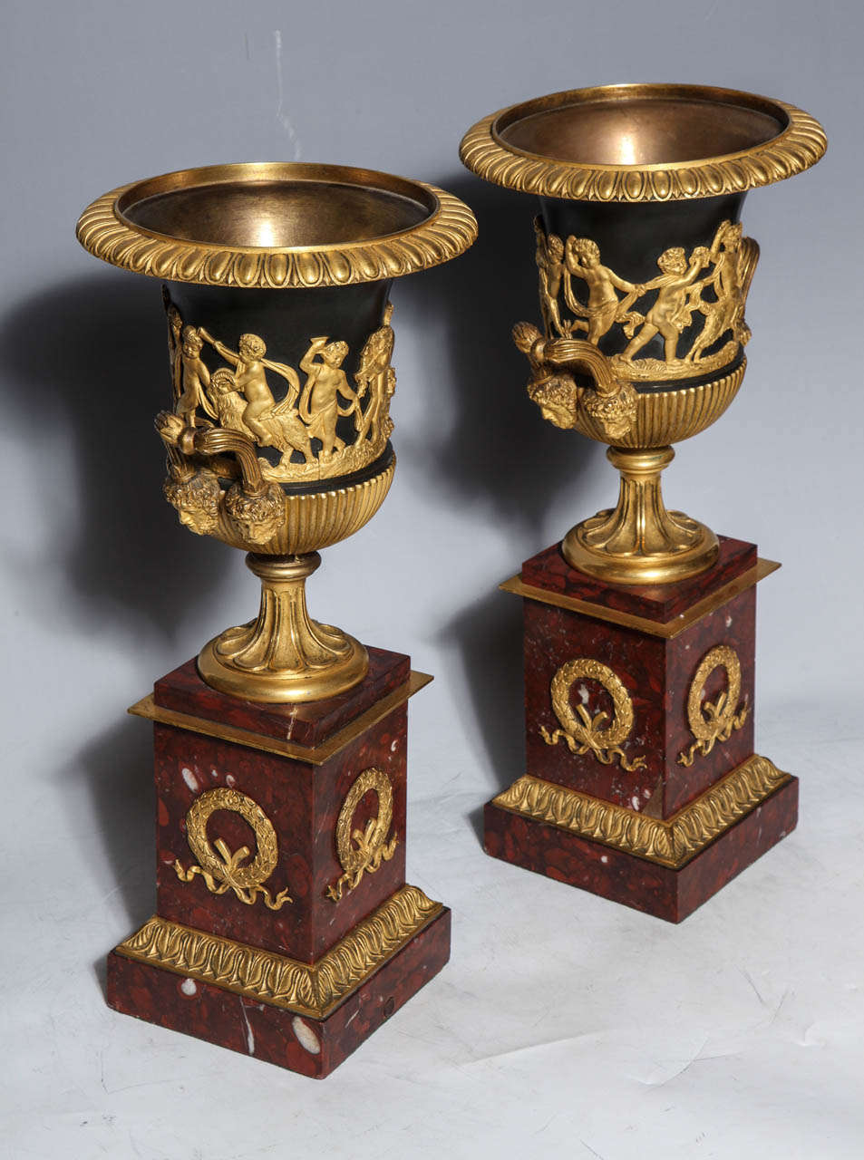A pair of neoclassical French Empire Period doré Bronze, patinated bronze and Rouge Campana shaped marble vases, circa 1820, with gilt bronze decorations of finely sculpted playful putti. Additionally, masks uphold the elegant flaring handles. The