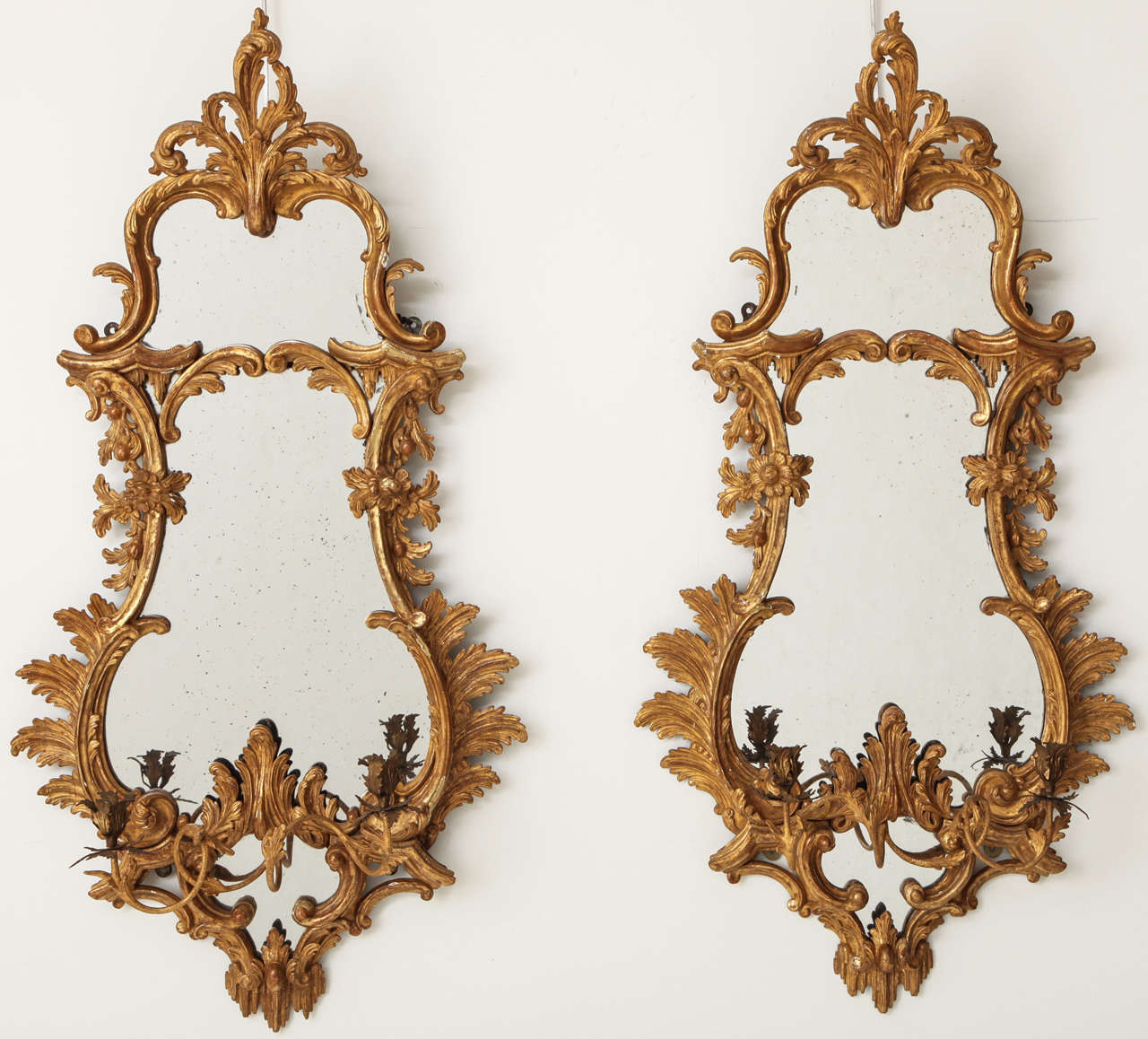 A pair of George III style carved giltwood girandole mirrors in the Rococo taste, each cartouche-form plate surmounted by a leafy crest above a  C-scroll and foliate garland border, the base centering a pair of spiraling vine candlearms.