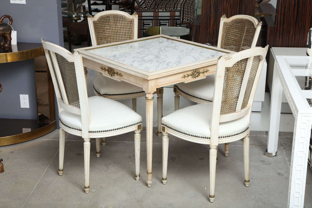 A neoclassically inspired game table with 4 chairs en suite.Taable and chairs are painted in a warm cream tone and accented with brass mounts. Chairs are covered in their original leather. Attibuted to Jansen