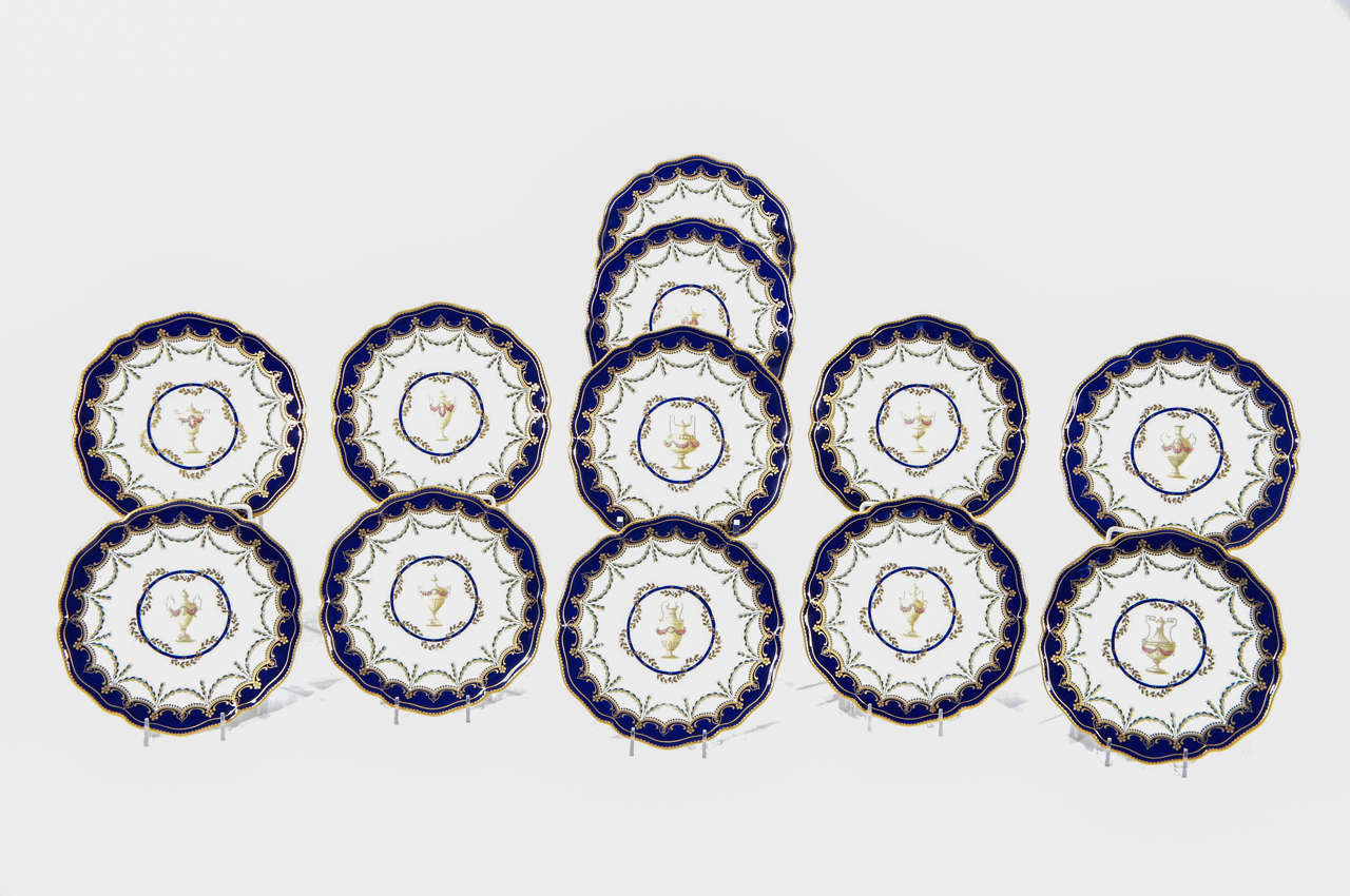 An unusual set of 12 Royal Crown Derby dessert plates featuring an elegant and restrained central subject of subtly different urns with floral decoration encircled with swags of golden leaves. The shaped rims are bordered with cobalt blue with
