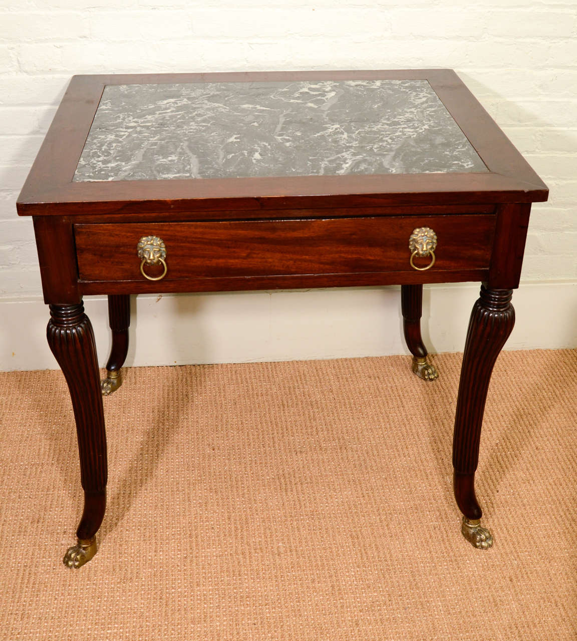 The top inlaid with a variegated grey marble top. The top over a frieze containing a single drawer with two lion's head gilt metal pulls. The table raised on four fluted and turned tapered sabre form legs ending in brass paw feet.