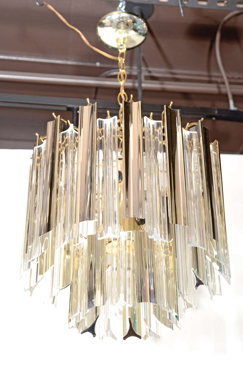 Modernist chandelier with brass frame and fittings. Chandelier has two tiers of alternating three sided lucite prisms. Lucite pendants are in clear and metallic moonglow, which reflect both brass and chrome hues.  This fixture is fitted with nine