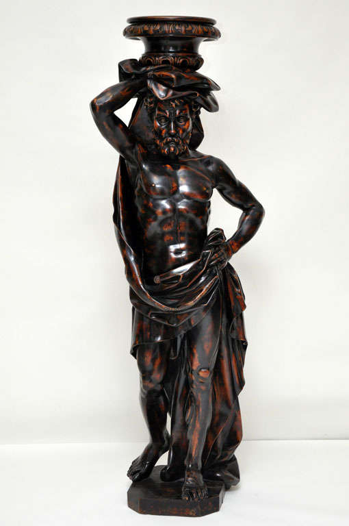 An intricately hand carved life-size bearded and barefoot Atlantid figural pedestal, cloak draped around his well defined body, the garment is gathered in his right hand above his head supporting a circular plinth shelf  with continuous acanthus