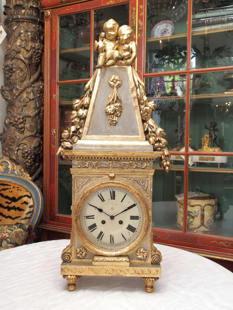 Italian Parcel Gilt and Painted Carved Wood Obelisk Form Clock with two putti and a bird and cat surmounting the obelisk on a rectangular base on acanthus feet.