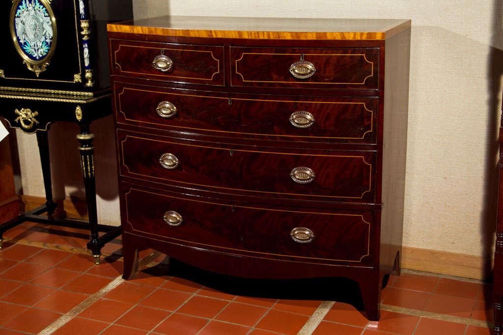 A fine English mahogany bow front chest with satinwood crossbanding to the top, with boxwood string inlays on the top and drawer fronts. A deep crotch mahogany is used on the drawer fronts, giving depth behind the inlay. The bowed apron and splayed