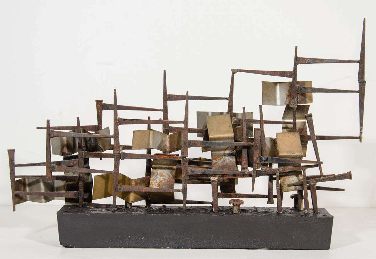Mid-century brutalist art sculpture comprised of mixed metals including flamed brass and copper metal squares and blackened and rusted horseshoe nails. The sculpture rests on elongated and narrow concrete base painted in black. The sculpture has an