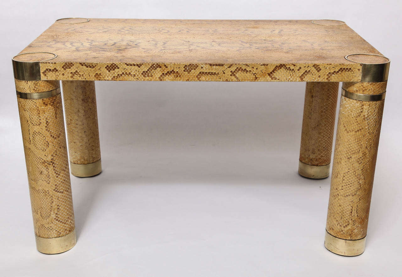 A 1970s architectural form table, following the style and designs of Karl Springer, covered in snakeskin against a wood base, standing on round legs, accented by brass mounts.