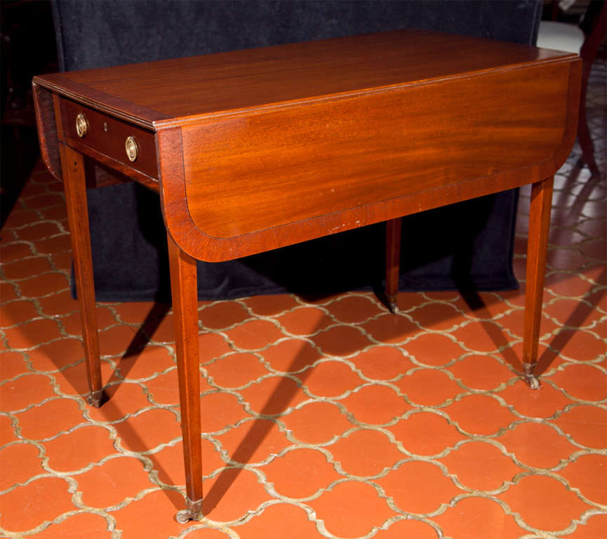 This large mahogany pembroke table is crossbanded in mahogany with an ebony stringing separating the band from the field. A triple reading finishes the edge of the table. A single oak-lined drawer with brass ring pulls provides generous storage