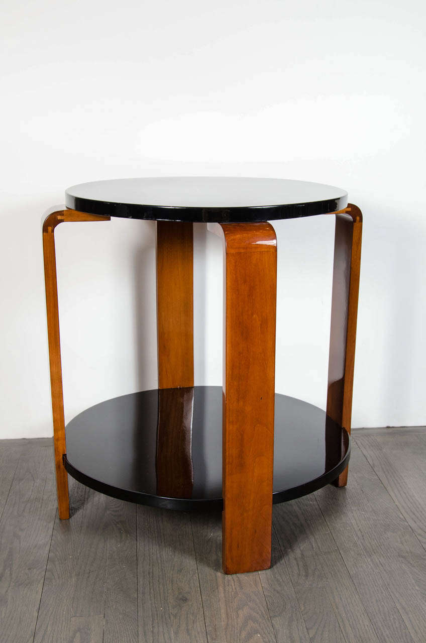 This elegant Machine Age Art Deco occasional table features two tiers, the bottom offering a slightly larger diameter than the top, in black lacquer that are connected by four rectangular walnut banded supports that culminate in streamlined tops.