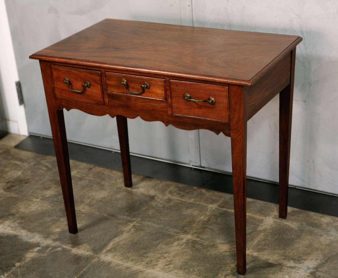 This George III mahogany side table has 3 small drawers. It stands on tapered legs. There is a scroll frieze front. The drawers are dovetailed with brass pulls. 