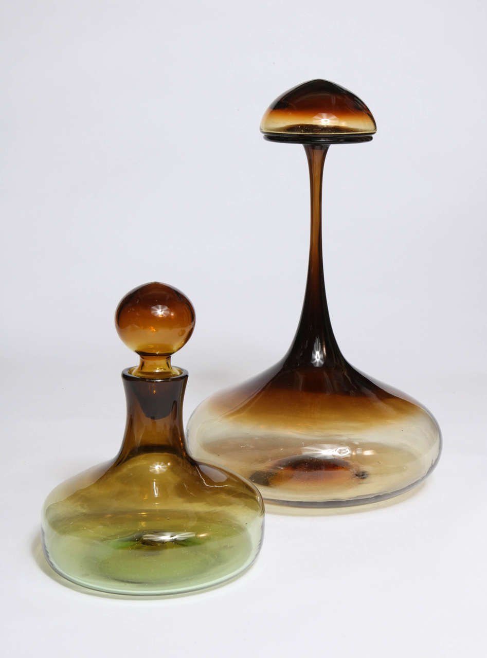 This decanter by Tom Connally for Greenwich Flint Craft features a heavy bottom, an extremely long neck, a flared top and a mushroom-shaped stopper. Burnt honey color with gold flecks. Model #1183, unsigned. 

The smaller decanter on left is sold.
