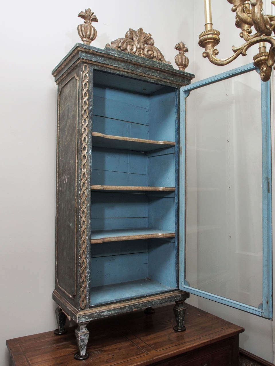 A petite, painted single door cabinet with silvered carving, topped with finials and a pierced cartouche.  The side panels, cornice and apron are molded, and the piece rests on tall, square, tapered feet.  Interiors shelves shaped and molded.