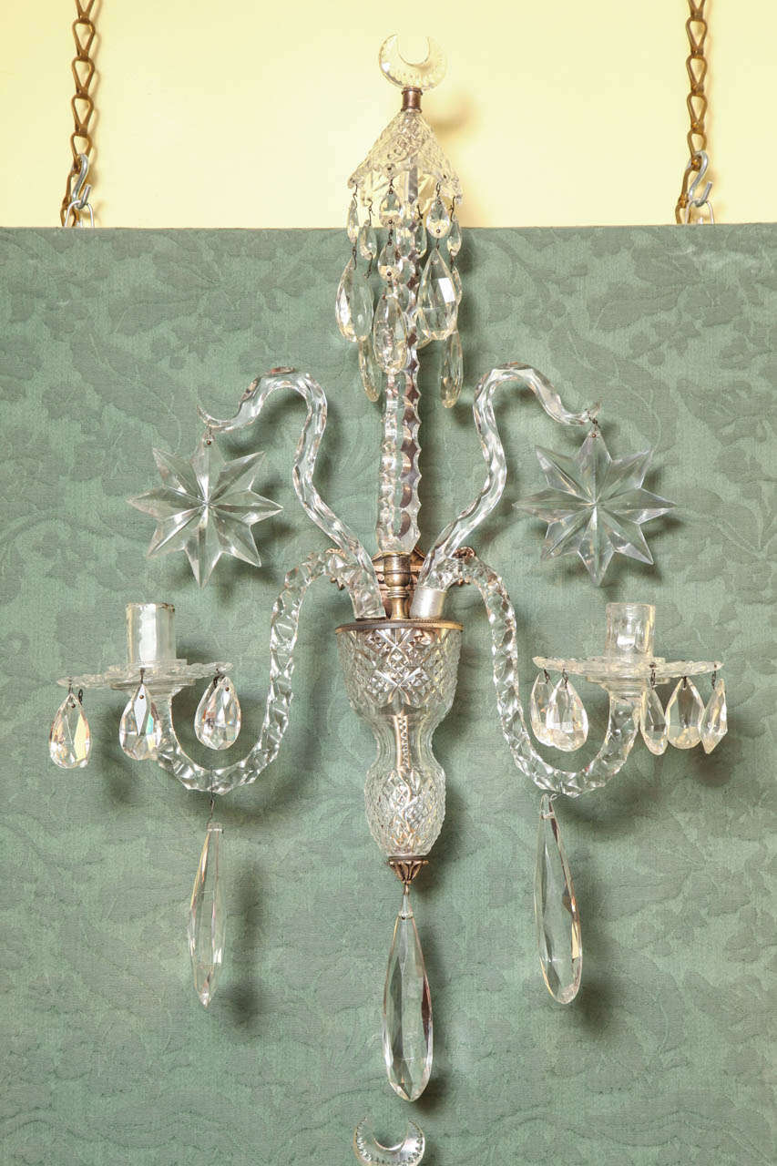 Fine pair of antique Edwardian cut crystal two-light wall lights, having a notched crescent finial above a triangular spire with faceted angles, with cut canopy with faceted drops and having two up scrolled arms with large star pendants above two