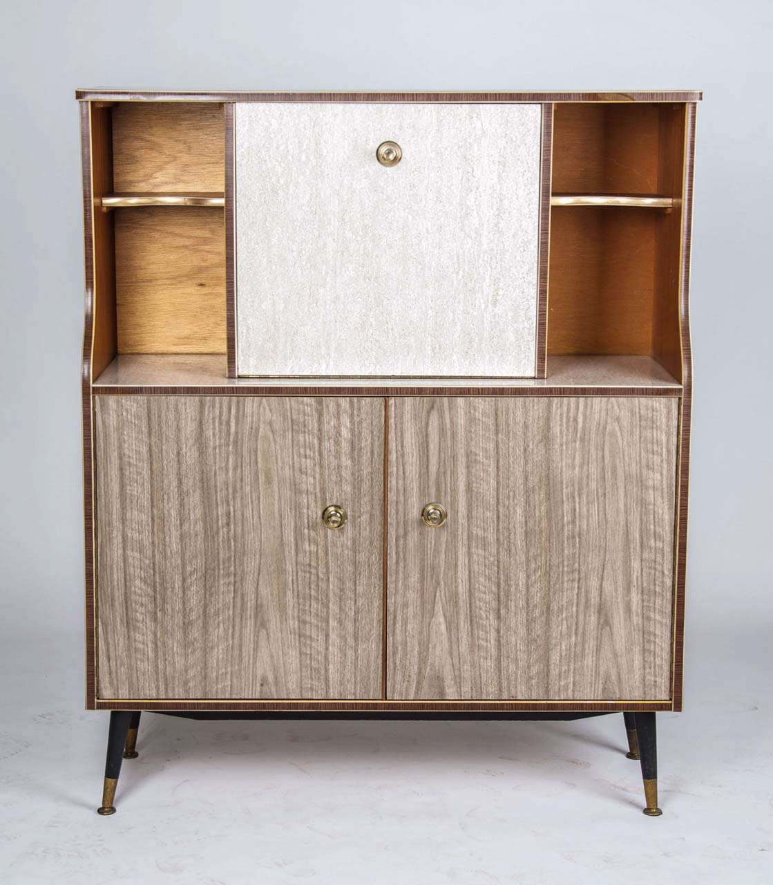 A 1960,s faux wood veneer sideboard in original condition, with one central pull down door cupboard door above a pair of pull open doors standing on ebonised feet capped in brass.